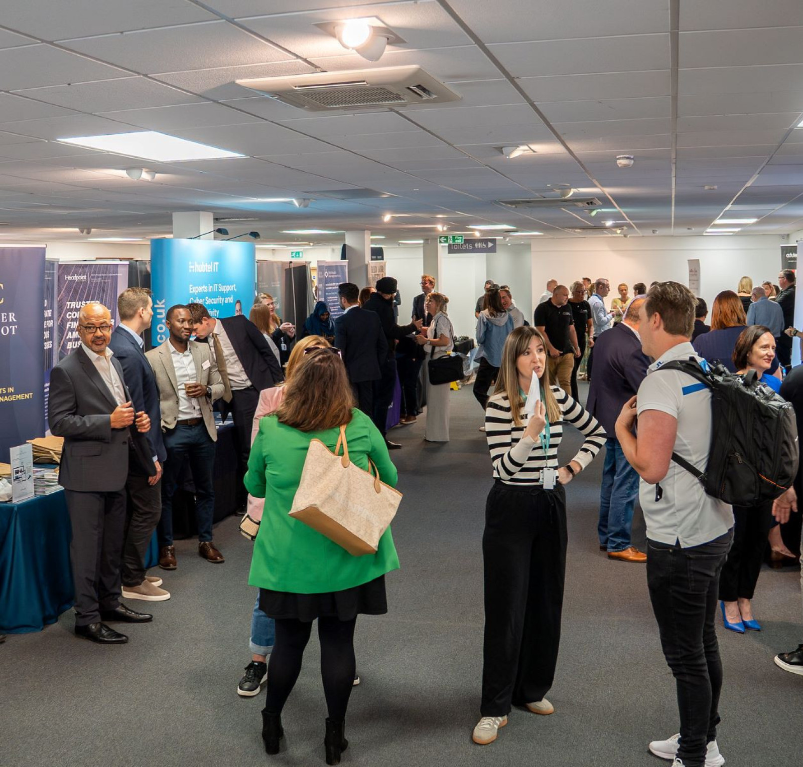 A bustling event space filled with people networking and engaging at various booths. Attendees are dressed in professional attire, with banners and tables showcasing different businesses and services.