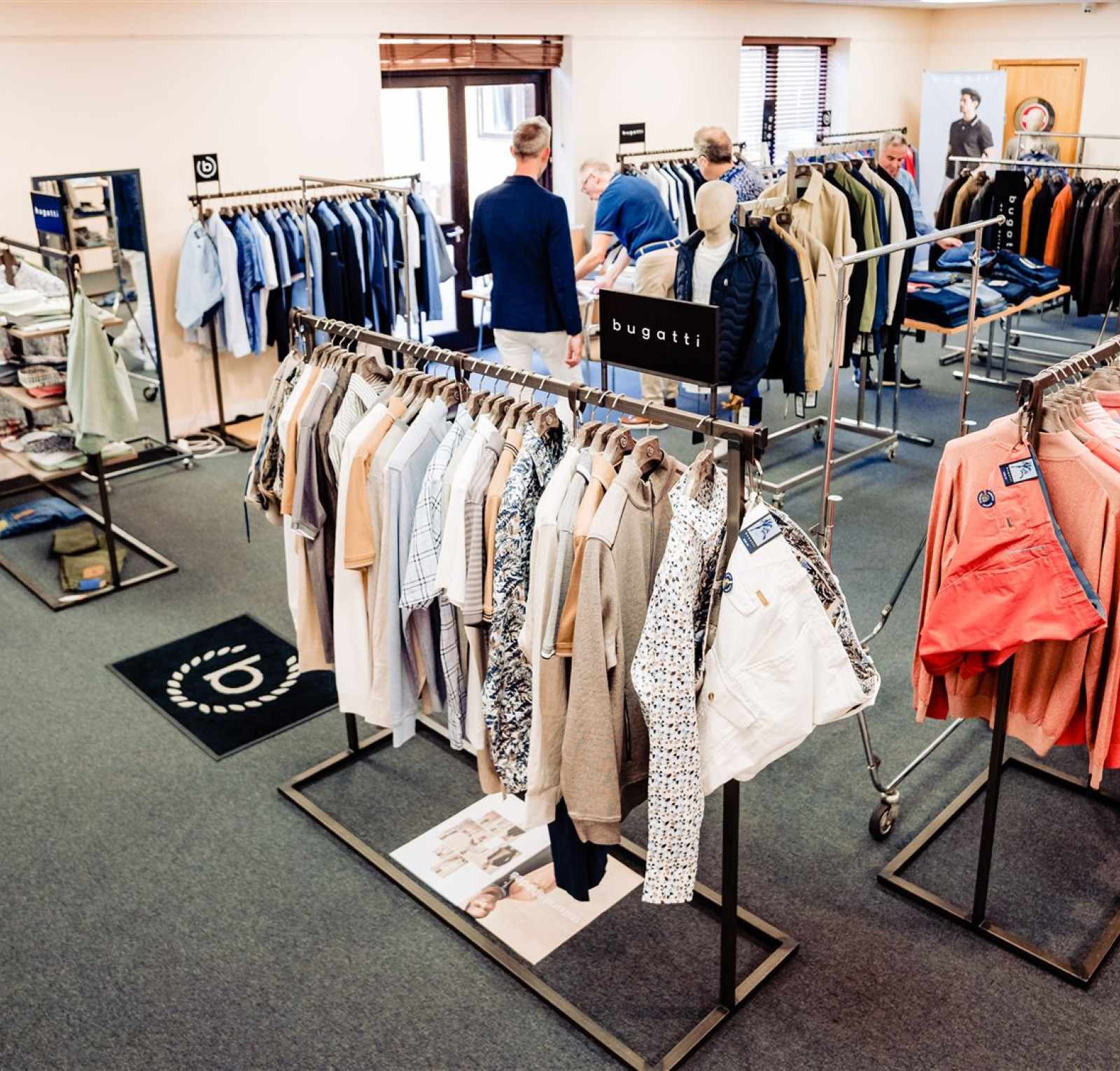 A clothing showroom featuring racks of various shirts, jackets, and pants. Several customers browse the items, while a TV screen displays images of clothing. The space has neutral-colored walls and a carpeted floor.