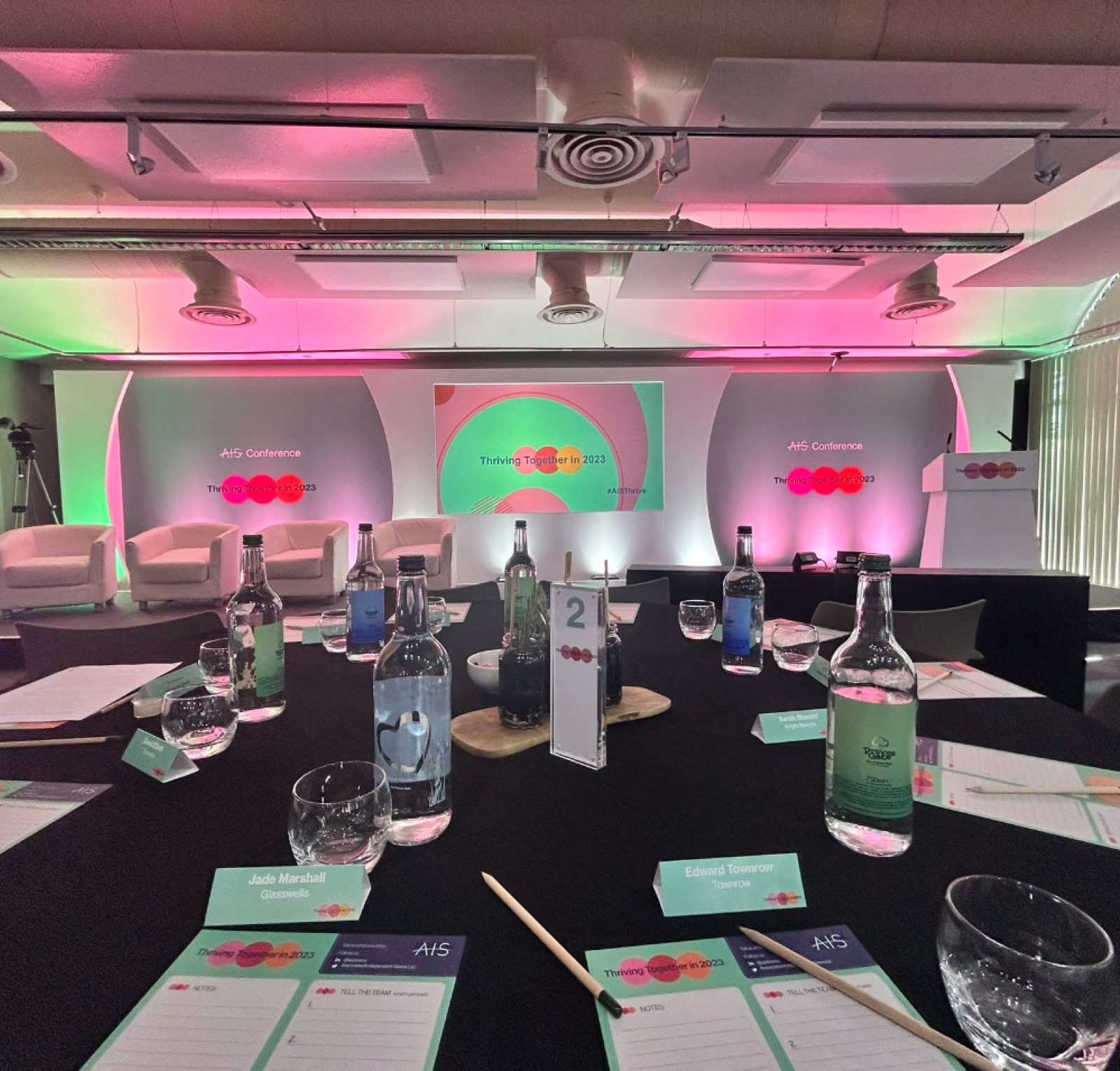 A conference room setup featuring a round table with notepads and pens. In the background, there is a colorful stage with lighting, equipped with a large screen and chairs arranged for a presentation.
