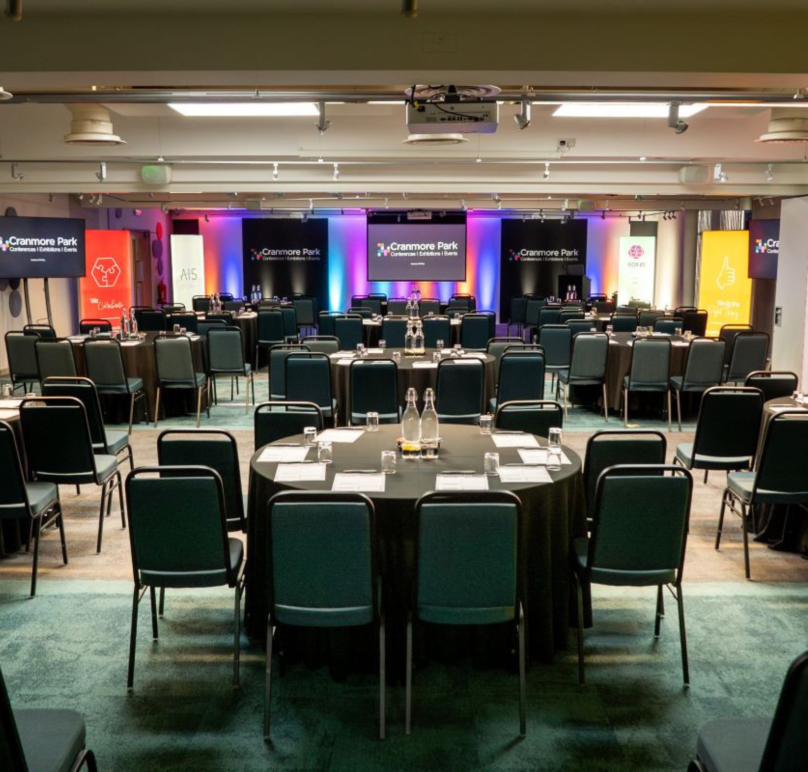 A spacious conference room set up for an event, featuring rows of round tables with black tablecloths and chairs. Colorful banners and screens are displayed at the front, with soft lighting enhancing the atmosphere.