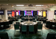 A spacious conference room set up for an event, featuring rows of round tables with black tablecloths and chairs. Colorful banners and screens are displayed at the front, with soft lighting enhancing the atmosphere.