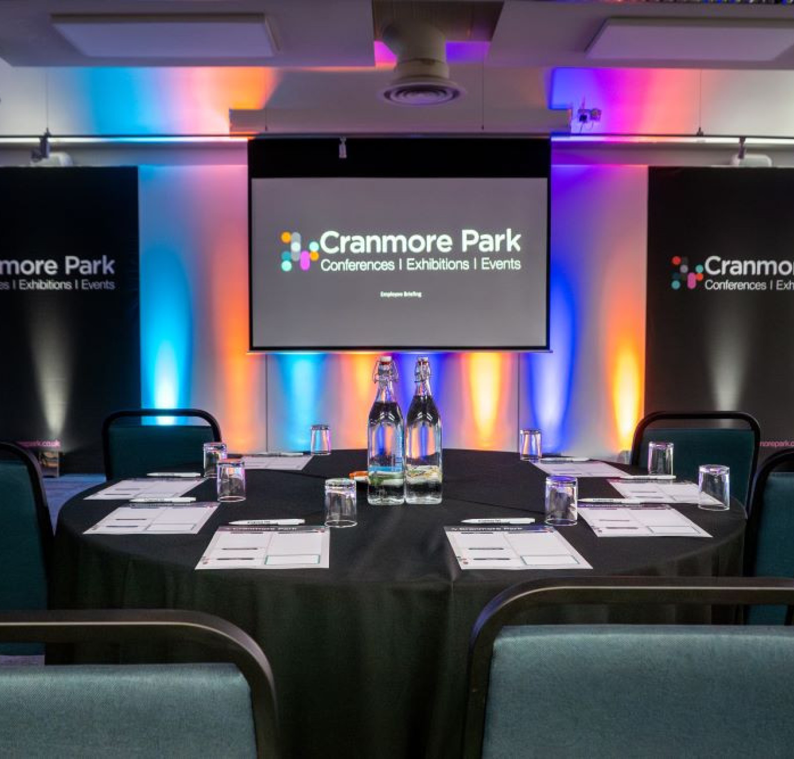 A conference setup with a round table covered in a black cloth, featuring bottles of water and glasses. Colorful ambient lighting enhances the space, with banners displaying 