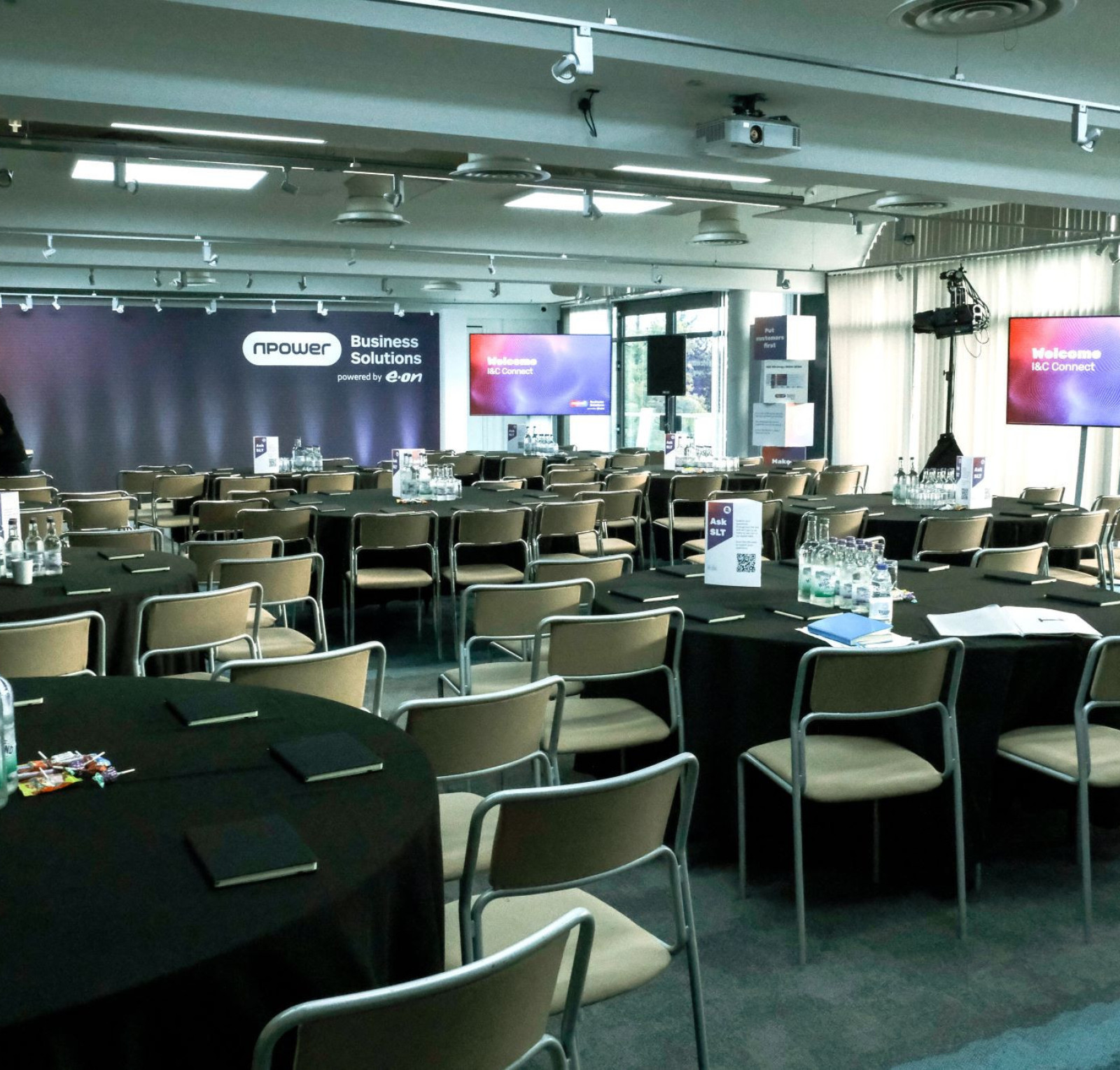 A conference room set up for an event, featuring rows of round tables with black tablecloths. Water bottles and notepads are on each table. A stage is in the background with large screens displaying a welcome message. The room is well-lit and has a modern design.