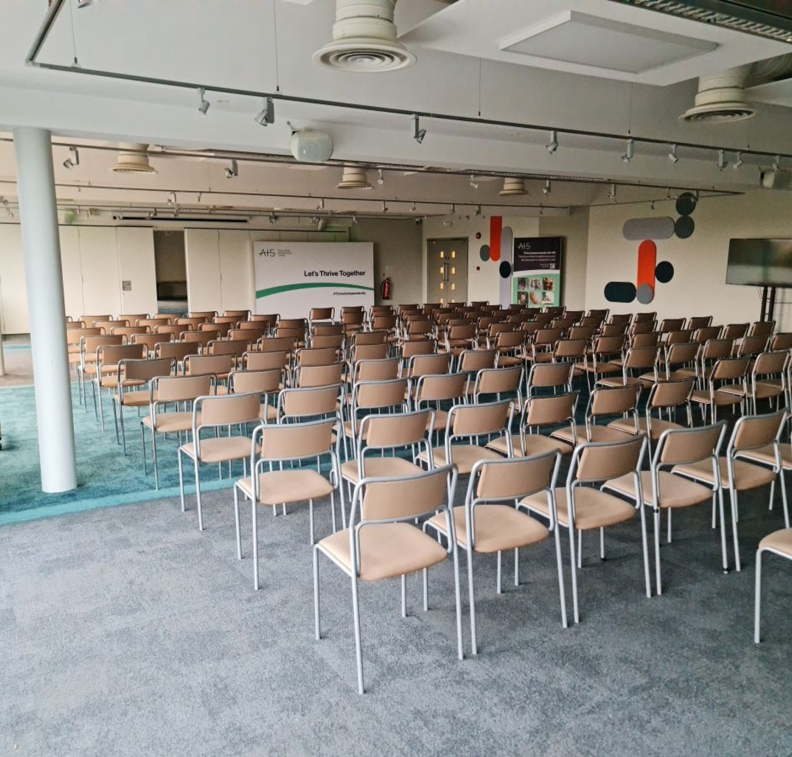 A spacious conference room with a row of beige chairs arranged in neat rows. The floor is carpeted in a muted color, and there are modern decor elements on the walls. A large screen is set up at the front of the room, and a banner in the background displays the words 