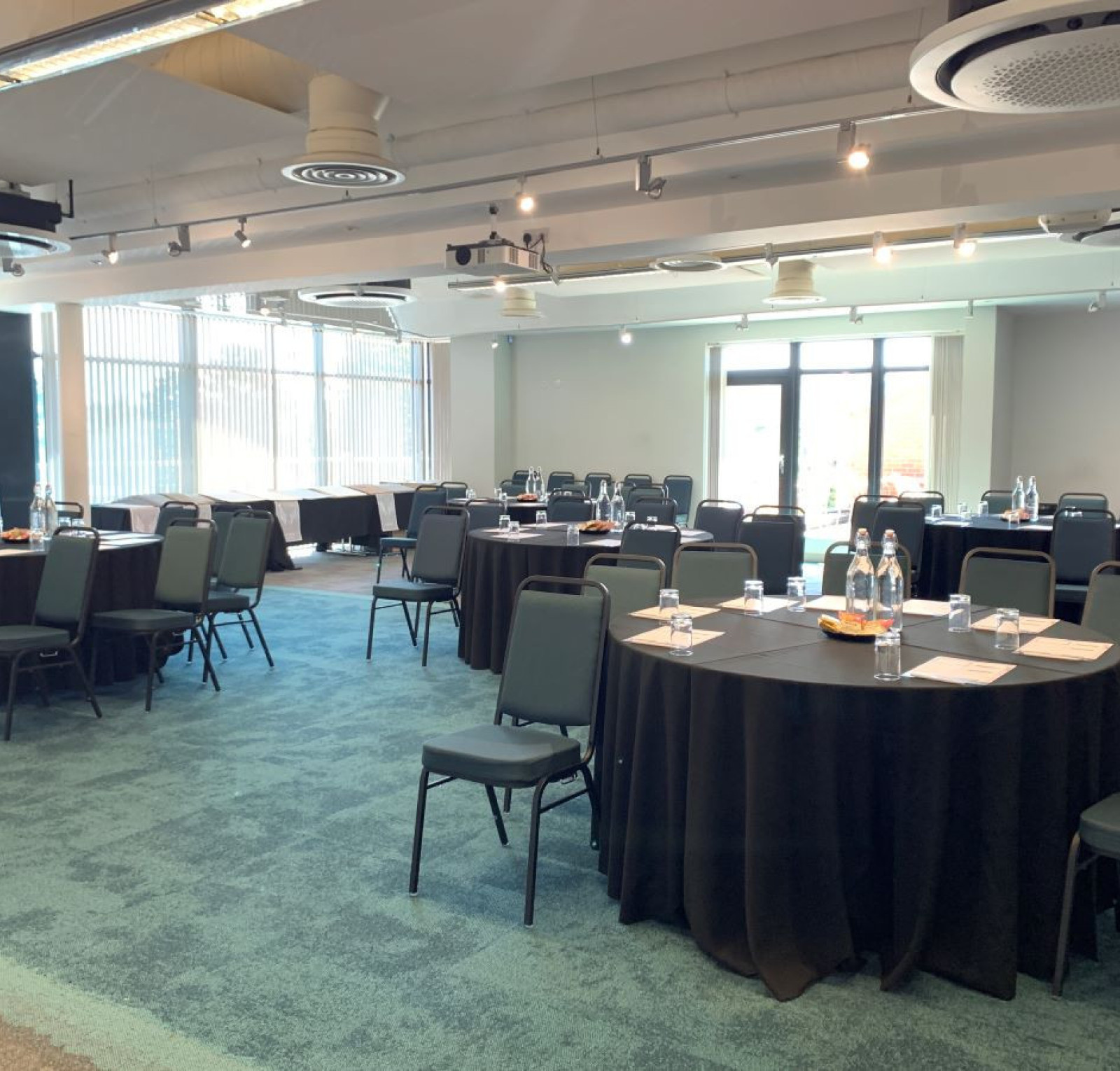 A spacious conference room set up with round tables covered in black tablecloths. Each table has water bottles and notepads, with green chairs arranged around them. Large windows let in natural light, illuminating the modern space.