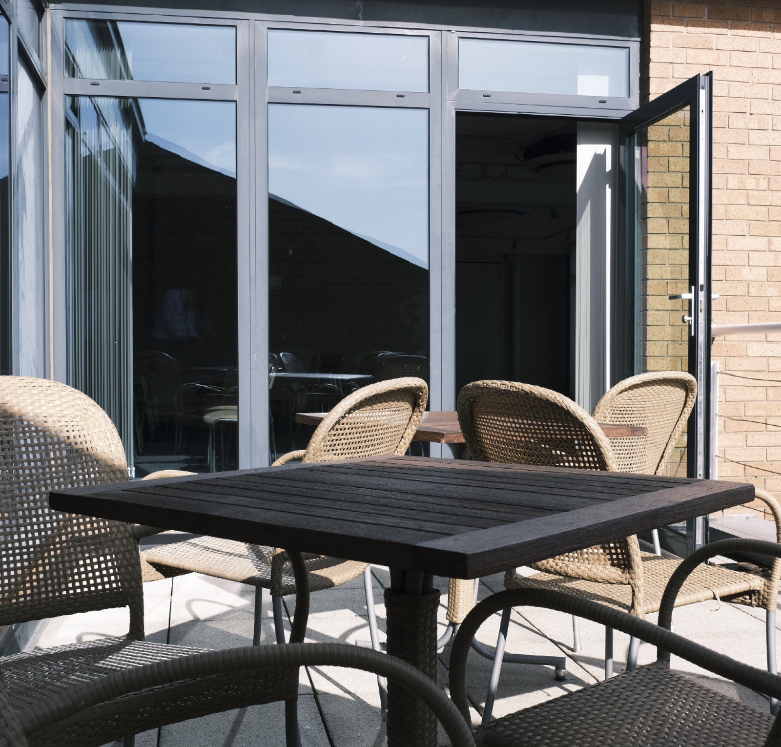 A cozy outdoor seating area featuring a wooden table surrounded by woven chairs. Large glass doors lead to an interior space, with sunlight illuminating the scene. A portion of a building with a sloped roof is visible in the background.
