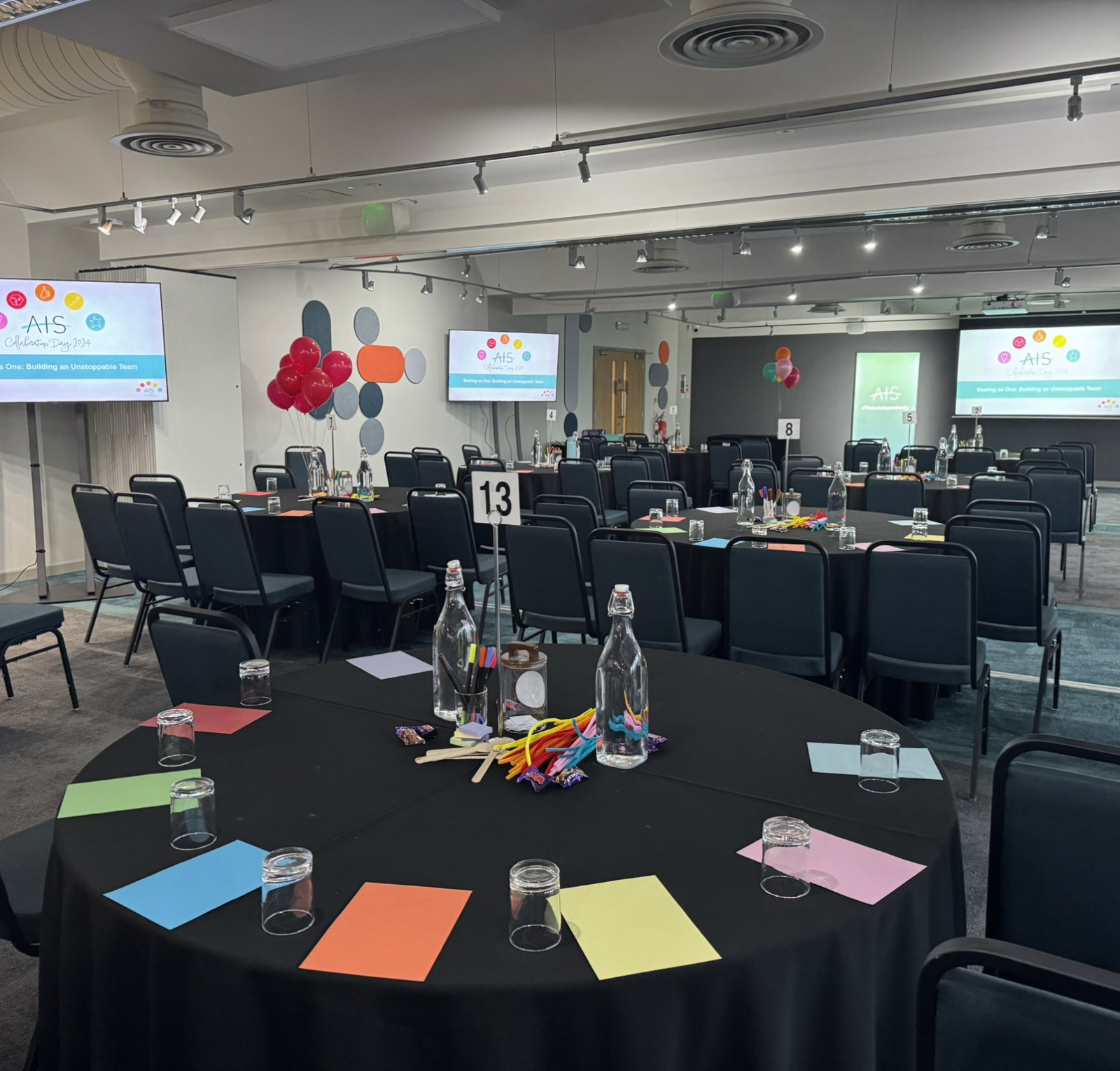 A conference room set up for an event, featuring several round tables with colorful place cards, water bottles, and stationery. Two large screens display the event logo, and balloons are decoratively arranged around the room.