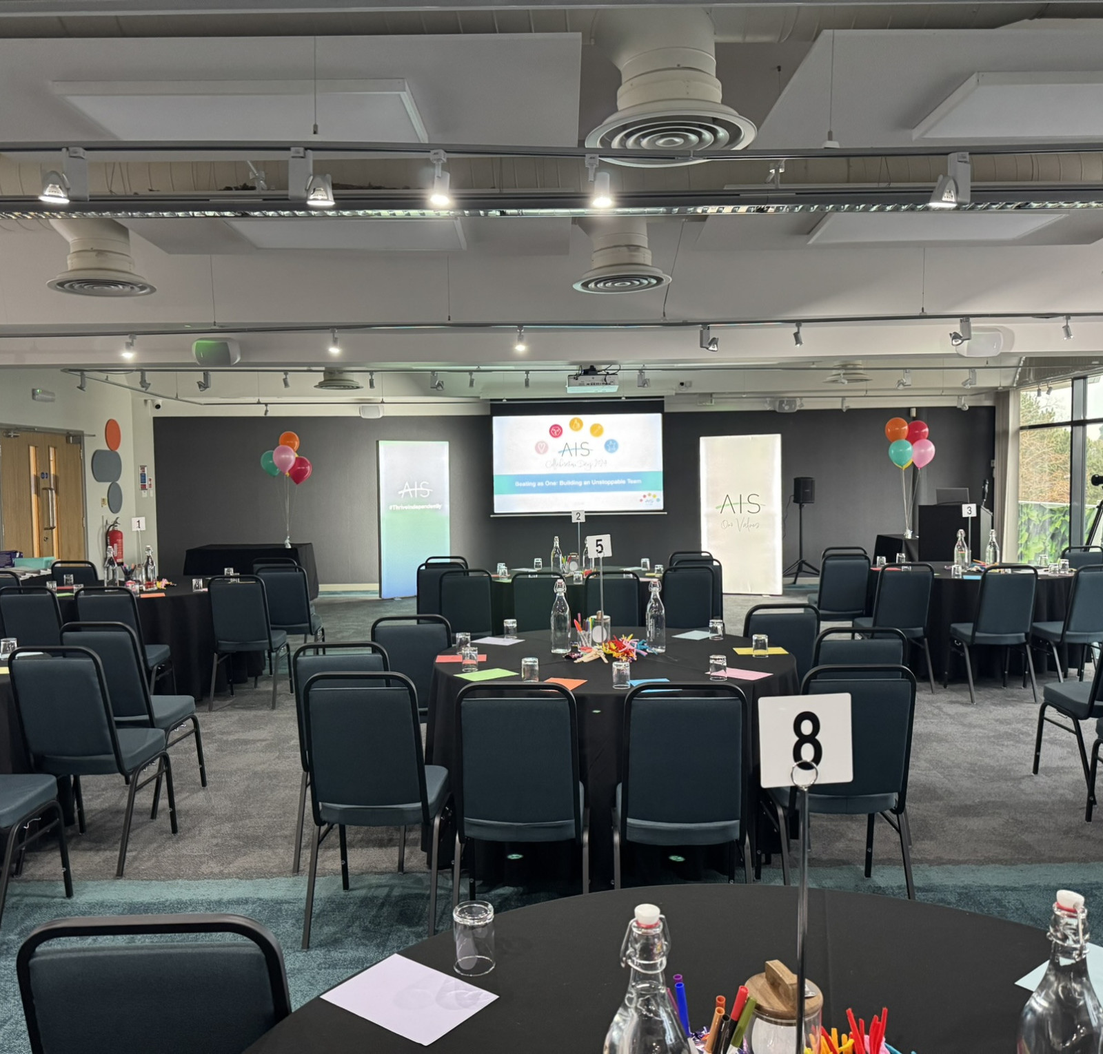 A large conference room set up for an event, with round tables covered in black cloths. Each table has water bottles, colorful pens, and name cards. Balloons decorate the space, and two large screens display presentation slides. The room is well-lit with windows showing greenery outside.