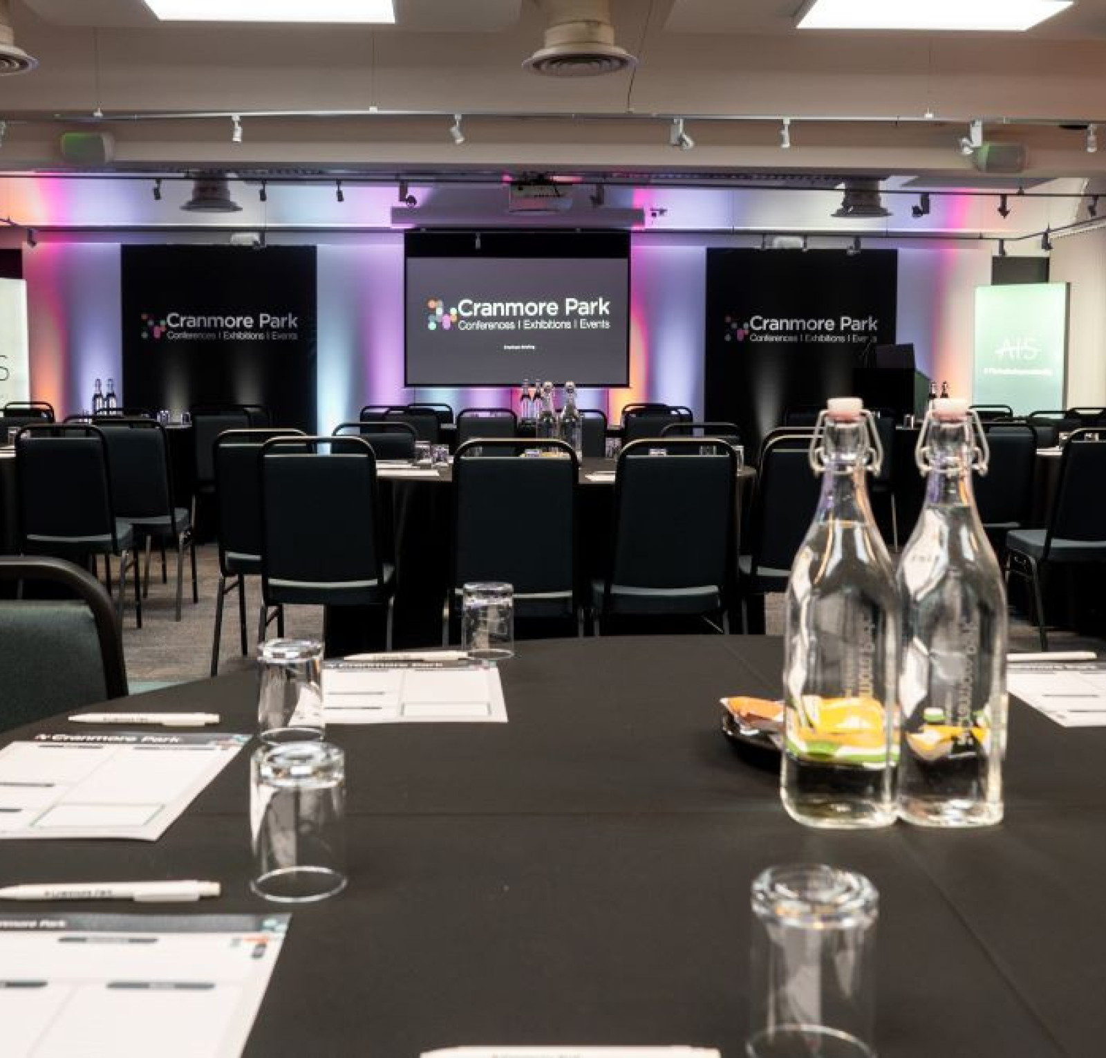 A conference room with round tables covered in black tablecloths, equipped with water bottles and glasses. Two screens at the front display the 