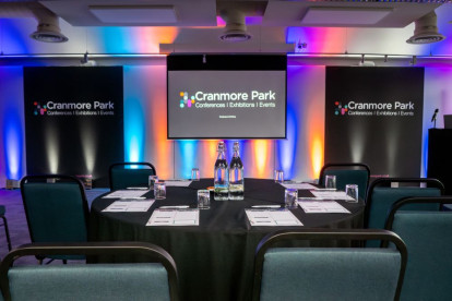 A well-set conference room with a round table covered in a black cloth. Water bottles and glasses are neatly arranged on the table, and presentation screens display the text 
