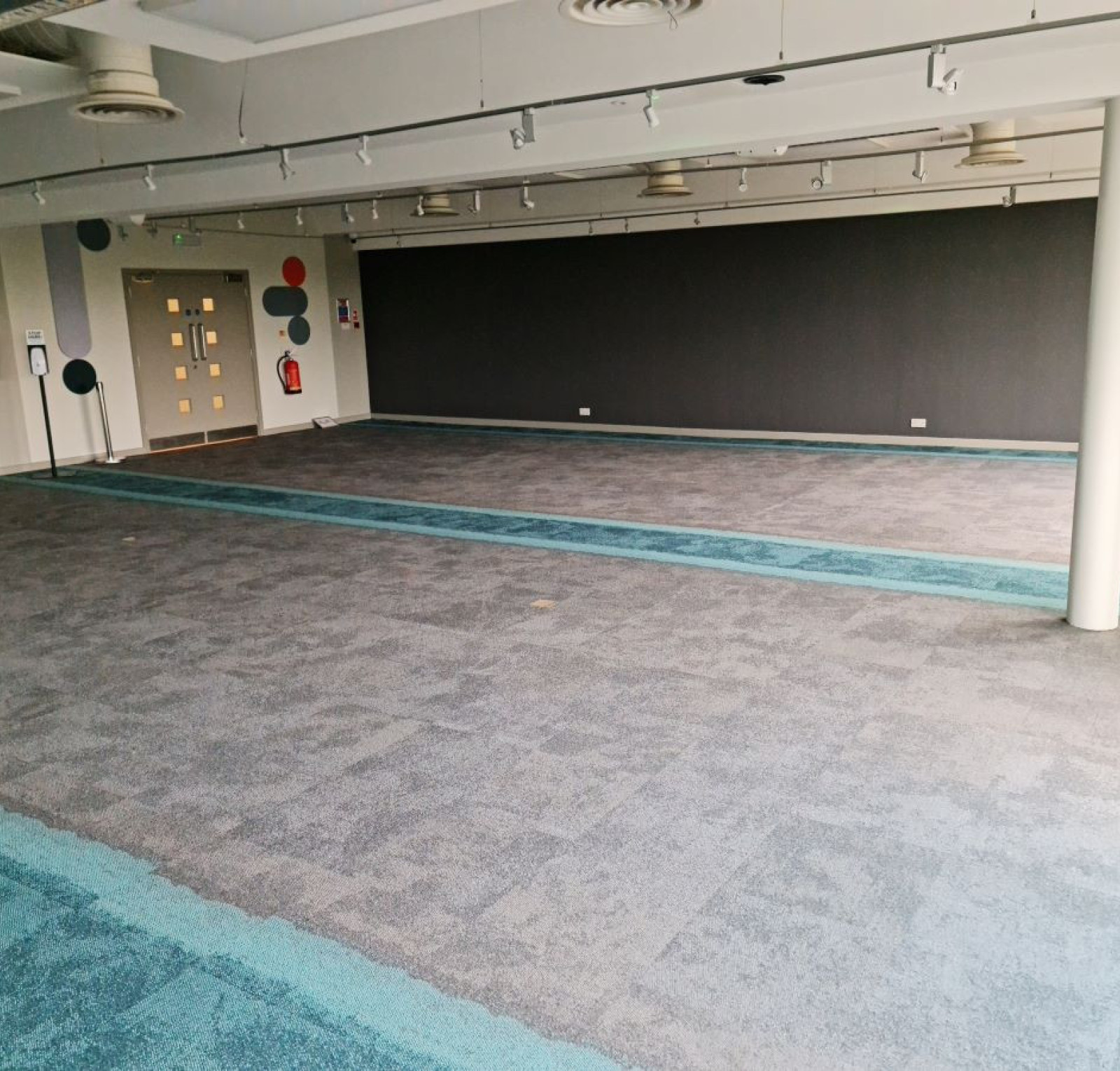 A spacious, empty interior room with gray carpet featuring turquoise accents. The walls are mostly dark, with a few colorful circular decorations. Bright overhead lights illuminate the area.