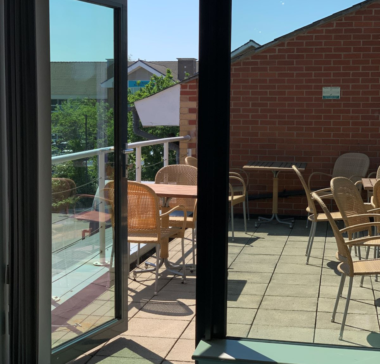 A sunny balcony with several wicker chairs and tables, viewed through an open glass door. The setting features a mix of brick and modern architecture in the background.