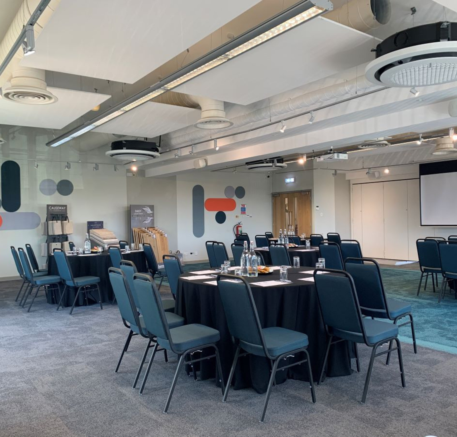 A conference room set up with multiple rows of dark blue chairs around tables covered in black cloth. The walls feature abstract designs in muted colors, and a projector screen is visible at the front of the room. Natural light illuminates the space.