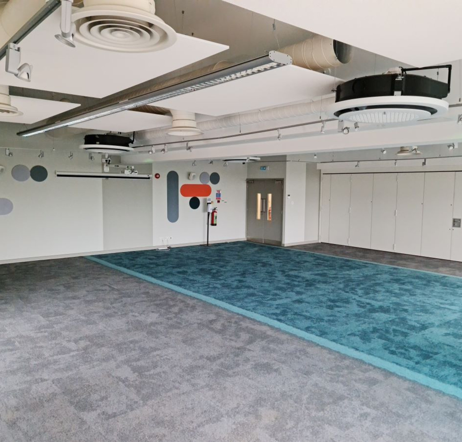 A spacious, modern room with gray carpet and a large teal carpet area. Walls feature abstract designs in orange, blue, and gray. There are ceiling-mounted lights and a projector system visible. Storage cabinets line one side of the room.