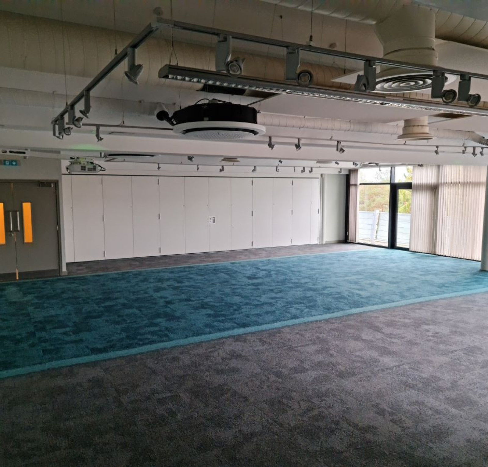 Empty meeting room with grey and teal carpeting, large windows allowing natural light, and storage cabinets along one wall. Ceiling features exposed ducts and lighting fixtures.