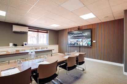 A conference room featuring a long table with chairs, water bottles, and stationary. A large screen displays a welcome message, and the walls have a modern design with wooden paneling. Natural light enters through a window.