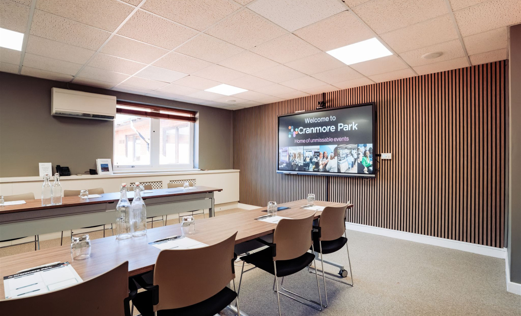 A conference room featuring a long table with chairs, water bottles, and stationary. A large screen displays a welcome message, and the walls have a modern design with wooden paneling. Natural light enters through a window.
