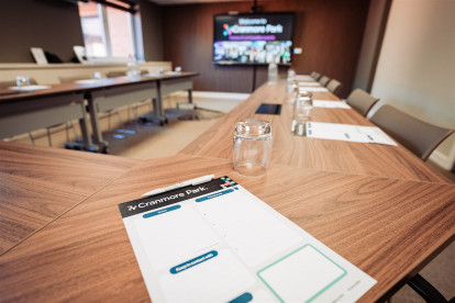 A conference room with a wooden table set for a meeting. Notepads are placed on the table, with a glass jar next to each one. A large screen displays the words 