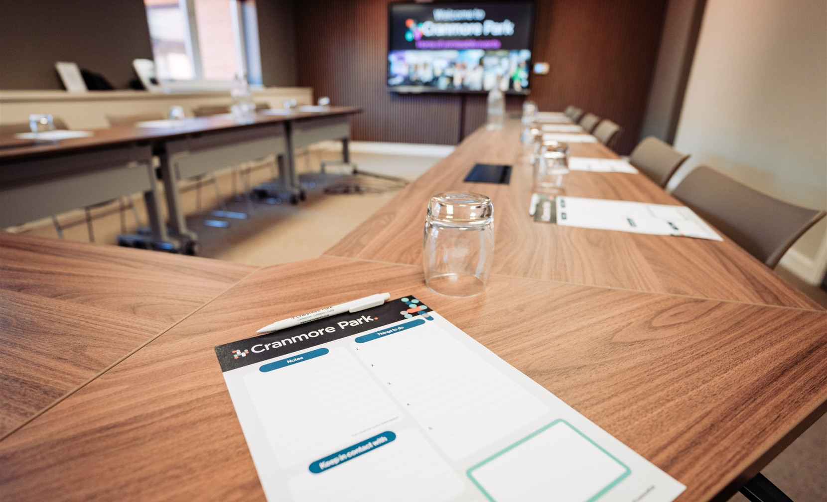 A conference room with a wooden table set for a meeting. Notepads are placed on the table, with a glass jar next to each one. A large screen displays the words 