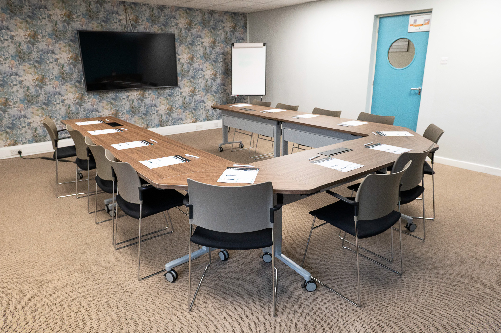 A modern conference room with a long, curved wooden table surrounded by gray chairs. There's a large screen on the wall, a flip chart stand, and a blue door with a circular window in the background. Floral wallpaper adds a decorative touch.