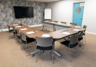 A modern conference room with a long, curved wooden table surrounded by gray chairs. There's a large screen on the wall, a flip chart stand, and a blue door with a circular window in the background. Floral wallpaper adds a decorative touch.