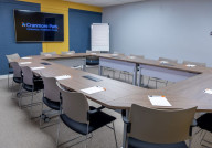 A modern conference room featuring a large, U-shaped wooden table surrounded by gray chairs. Each seat has a notepad and pen, and a projector screen displays the 
