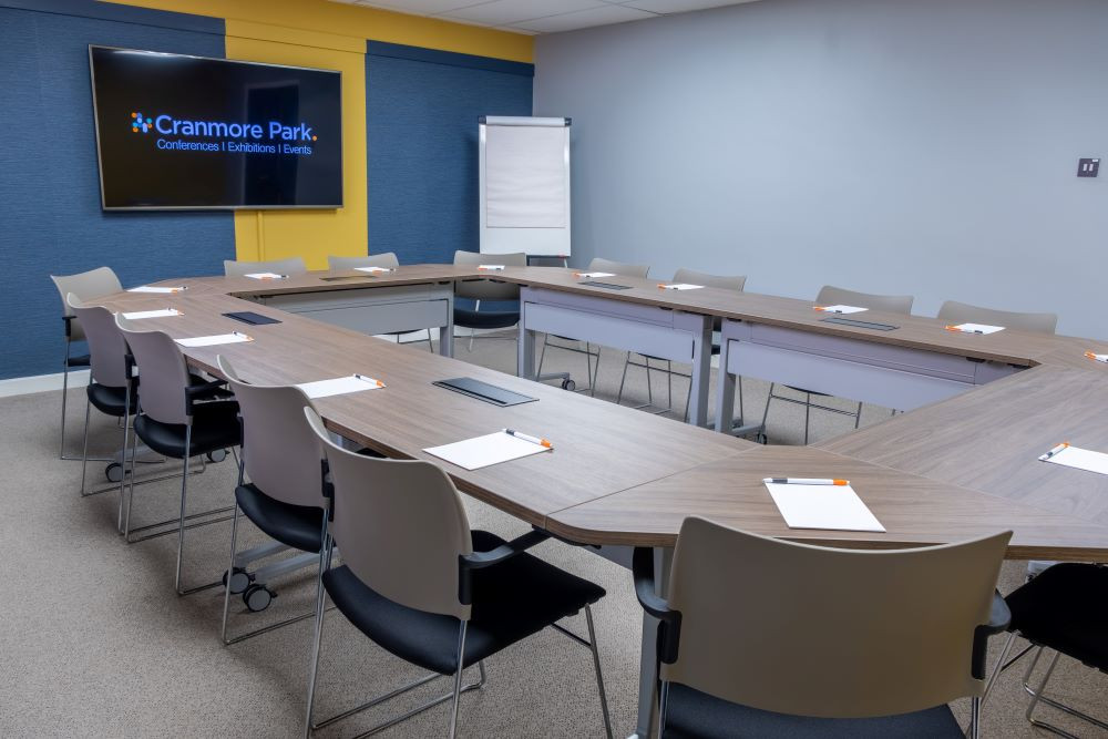 A modern conference room featuring a large, U-shaped wooden table surrounded by gray chairs. Each seat has a notepad and pen, and a projector screen displays the 