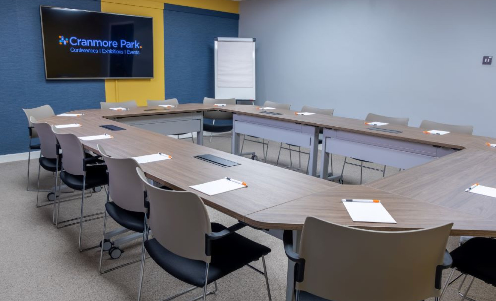 A modern conference room featuring a large, U-shaped wooden table surrounded by gray chairs. Each seat has a notepad and pen, and a projector screen displays the 