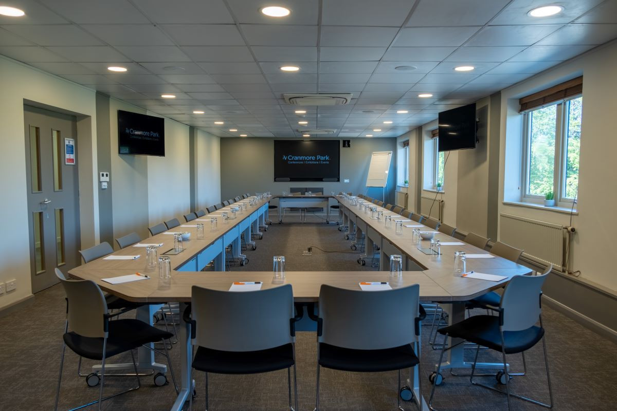 A modern conference room set up with a long, rectangular table surrounded by chairs. Water glasses and notepads are placed at each seat. Large windows allow natural light, and there are screens mounted on the walls for presentations.