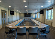A modern conference room set up with a long, rectangular table surrounded by chairs. Water glasses and notepads are placed at each seat. Large windows allow natural light, and there are screens mounted on the walls for presentations.