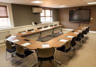 A conference room with a U-shaped table arrangement, equipped with chairs around it. The room features a large screen on one wall and windows with blinds. Notepads and pens are placed at each seat, and the walls have a modern design with wooden accents.