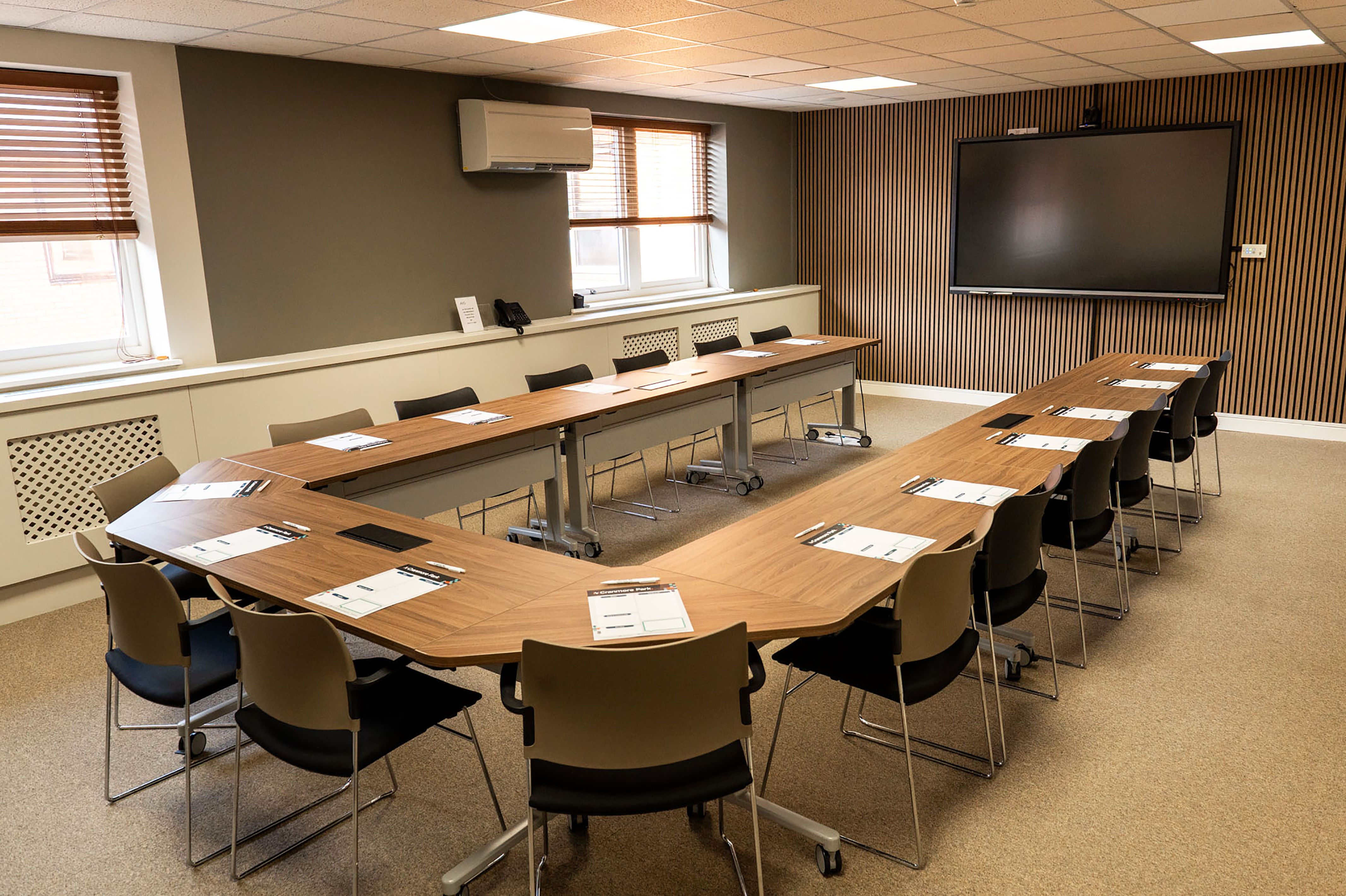 A conference room with a U-shaped table arrangement, equipped with chairs around it. The room features a large screen on one wall and windows with blinds. Notepads and pens are placed at each seat, and the walls have a modern design with wooden accents.