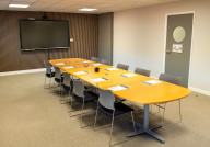 A modern conference room featuring a long wooden table surrounded by gray chairs. A large screen is mounted on the wall, and there's a door with a circular window. The floor is carpeted and the walls are partially paneled.