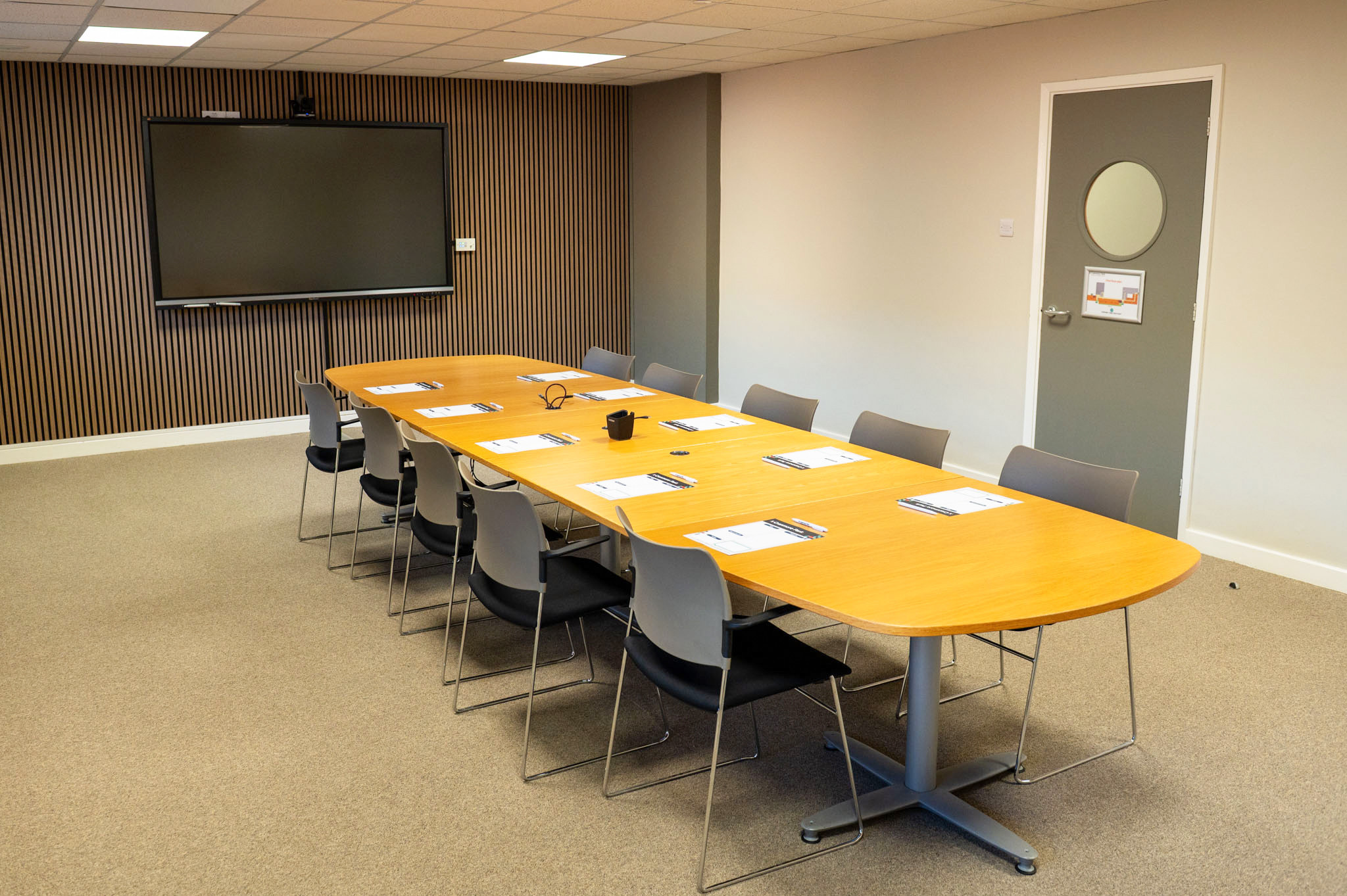 A modern conference room featuring a long wooden table surrounded by gray chairs. A large screen is mounted on the wall, and there's a door with a circular window. The floor is carpeted and the walls are partially paneled.
