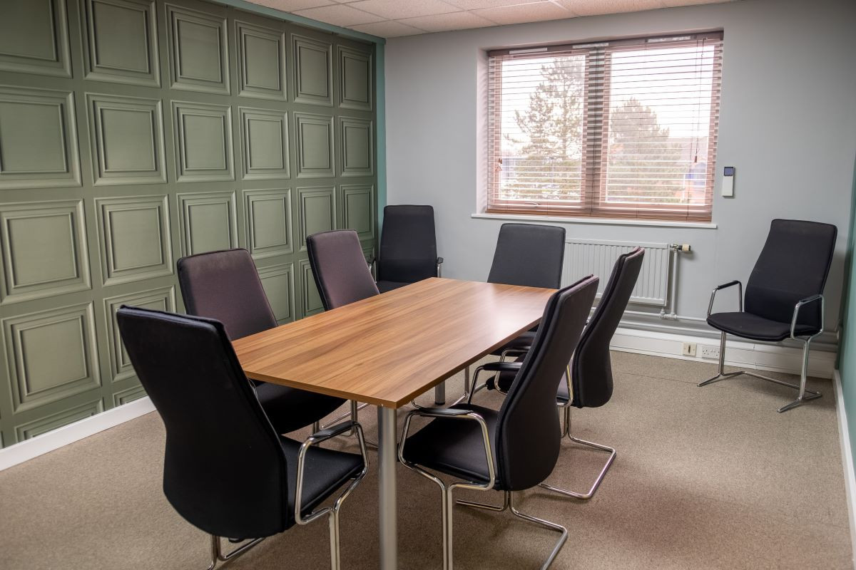 A modern conference room featuring a wooden table surrounded by six black chairs, with paneled green walls and a large window letting in natural light.