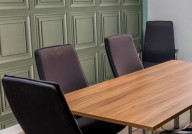 A modern conference room featuring a wooden table surrounded by six black chairs, with paneled green walls and a large window letting in natural light.