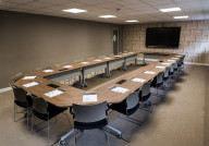 A modern conference room featuring a large, U-shaped table surrounded by multiple chairs. Each place at the table is set with notepads and pens. A flat-screen monitor is mounted on the wall, and the room has beige walls with a textured design.