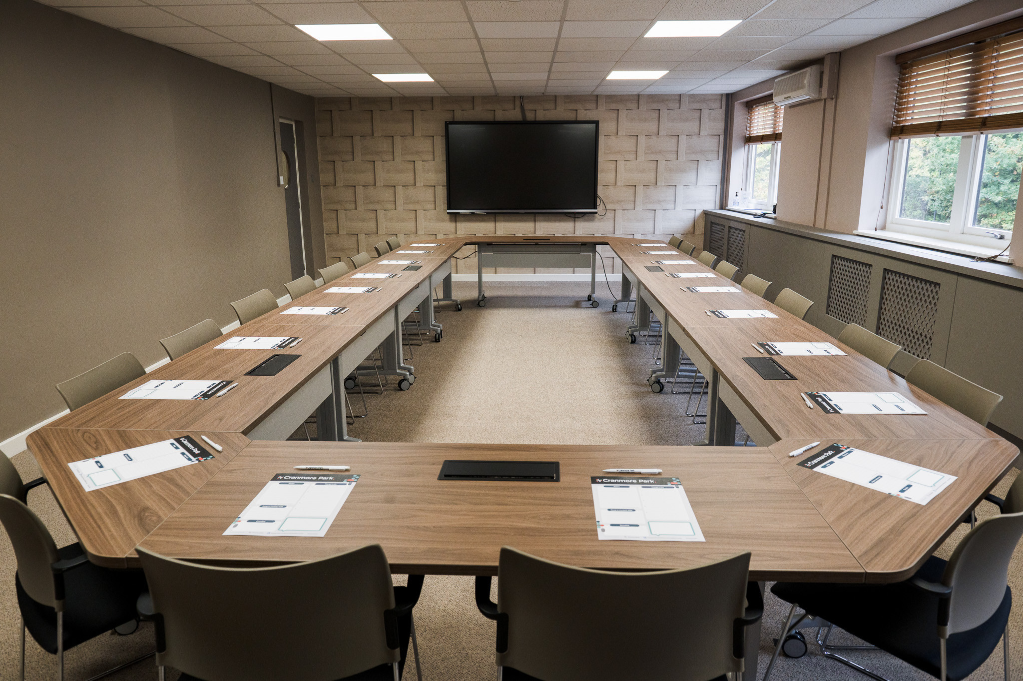 A modern conference room featuring a rectangular arrangement of multiple tables with chairs. Each table has notepads and pens placed on top. A large screen is mounted on the wall at one end of the room, and the walls are a neutral color with natural light coming through the windows.