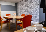 A meeting room featuring a round wooden table with four orange chairs. A wall adorned with black and white cityscape wallpaper is in the background. There are two bottles of water and glasses on the table, along with notepads and pens. Two white coffee cups are placed on a side table.
