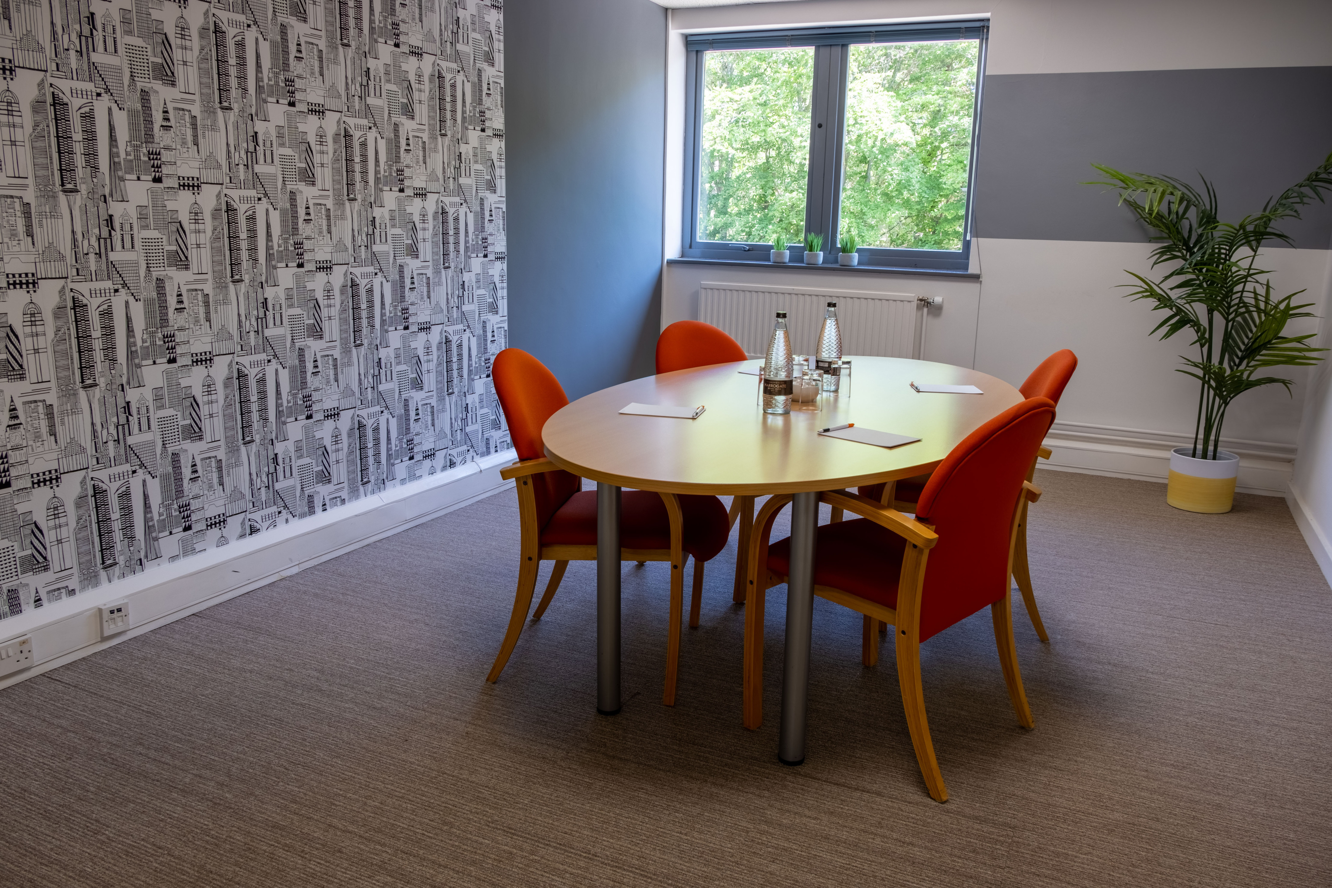 A bright meeting room featuring a round table surrounded by four orange chairs. A bottle of water and notepads are on the table. A large window offers a view of greenery outside, and a potted plant adds a touch of decor. The walls are decorated with a black and white graphic pattern.