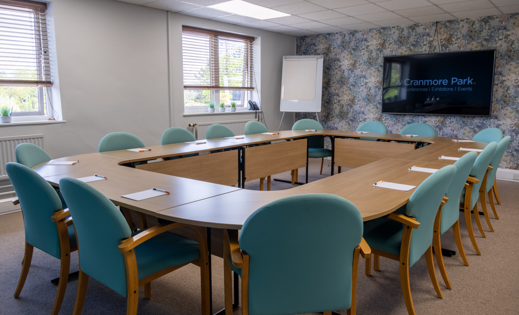 A well-lit conference room with a large, oval wooden table surrounded by teal chairs. The walls are partially covered with floral wallpaper, and a large screen displays the 