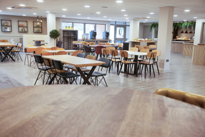 A spacious dining area featuring multiple wooden tables and chairs in various styles. The room is well-lit, with large windows and plants adding greenery. A buffet area is visible in the background, and modern decor enhances the ambiance.