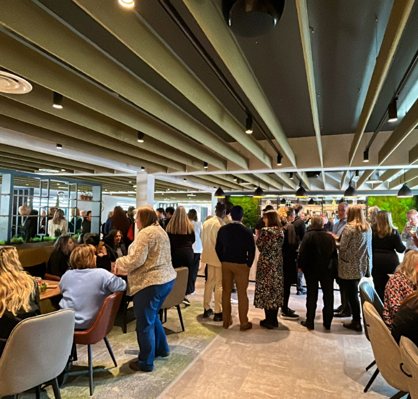 A crowded modern dining area filled with people engaged in conversation. The decor features green accents and wooden ceiling beams, creating a lively atmosphere. Some attendees are seated at tables while others stand and mingle.