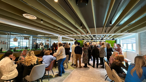 A crowded modern dining area filled with people engaged in conversation. The decor features green accents and wooden ceiling beams, creating a lively atmosphere. Some attendees are seated at tables while others stand and mingle.