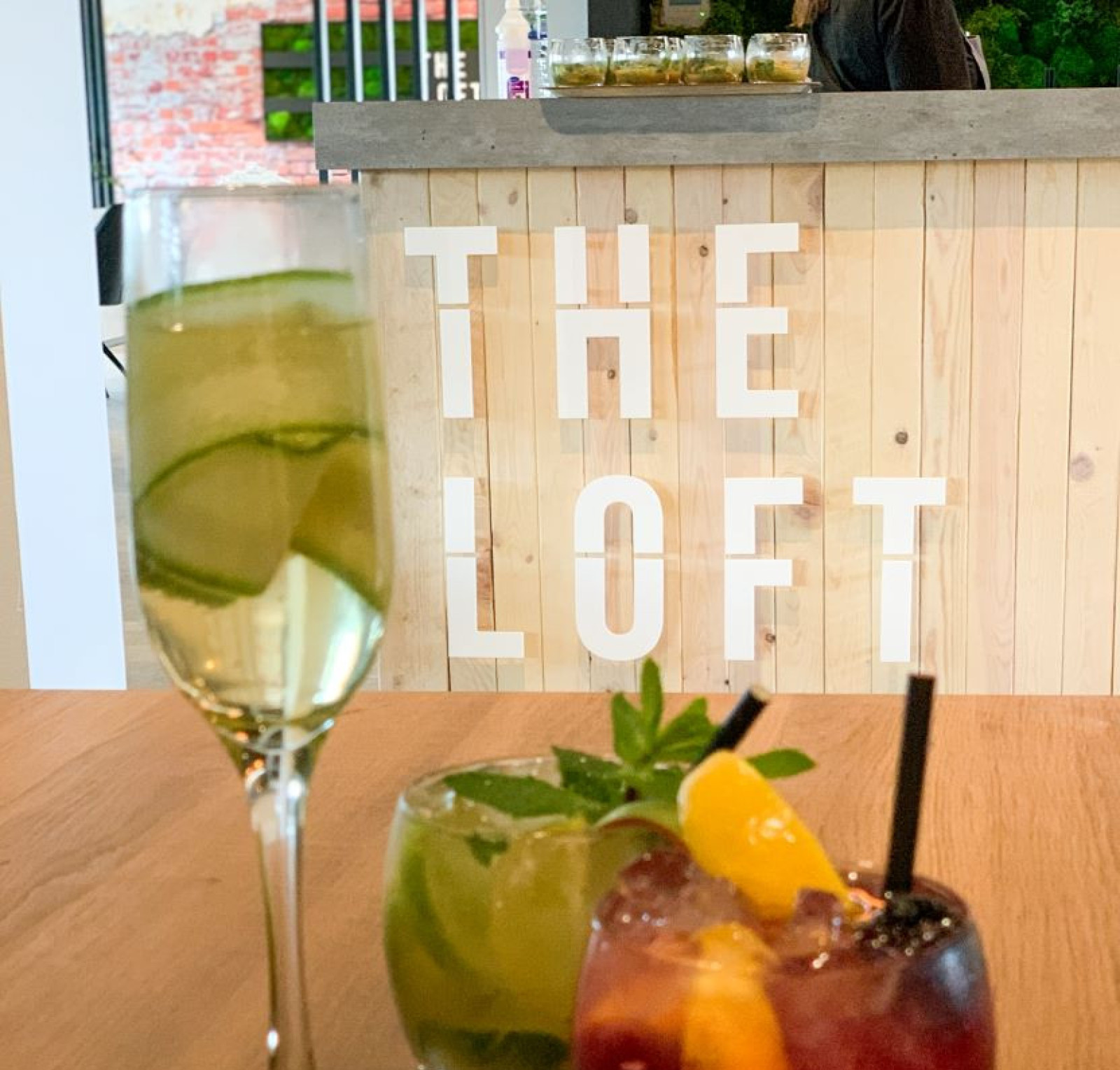 Three cocktails are arranged on a wooden table. On the left, a tall glass filled with a green drink and cucumber slices. In the center, a short glass contains a green cocktail, garnished with mint leaves. On the right, a short glass holds a dark red drink with an orange slice and a cherry on top. Behind them, the words 