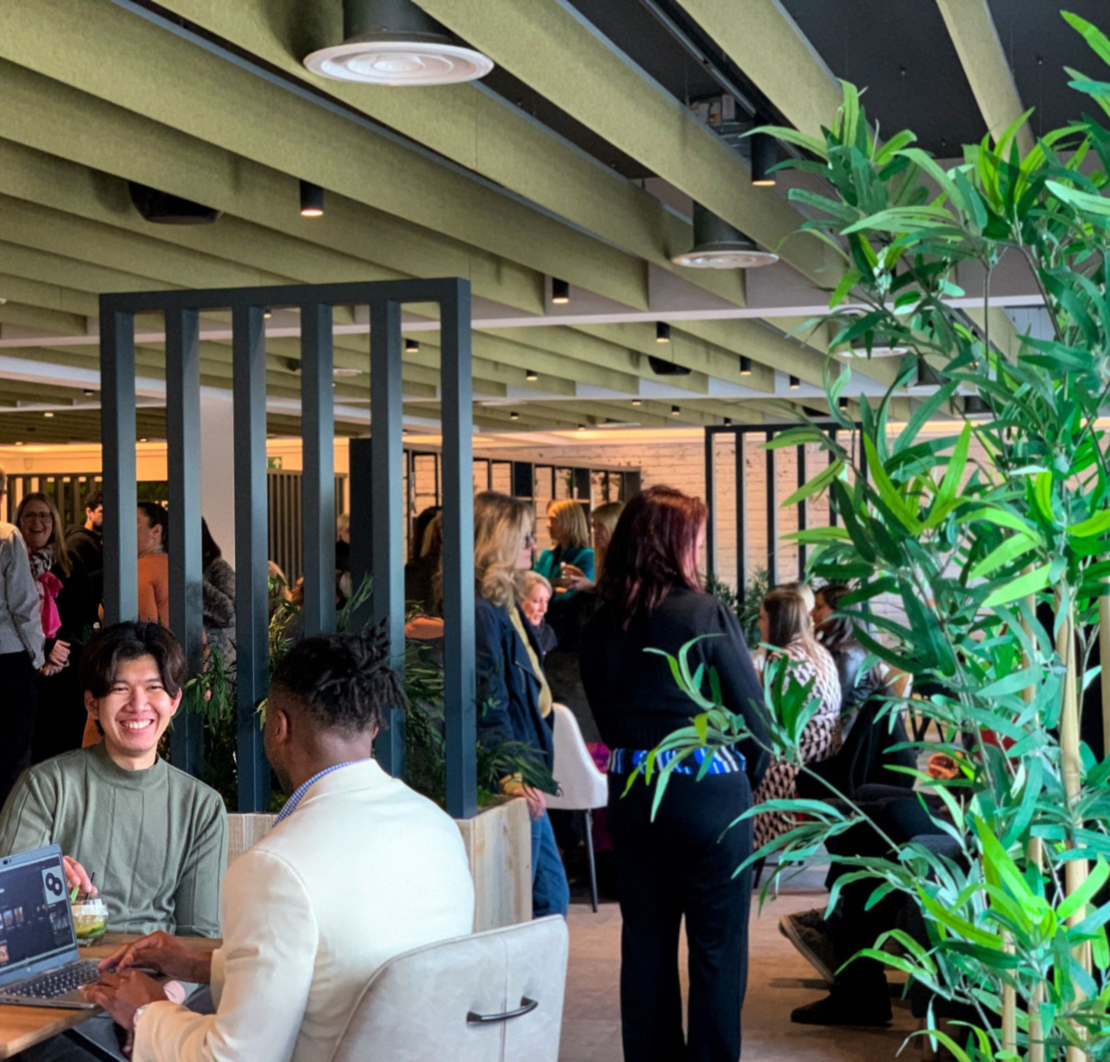 A busy café with people socializing. A man in a green shirt smiles while sitting at a table with a laptop, and another man in a white blazer is beside him. Lush green plants decorate the space, creating a vibrant atmosphere.