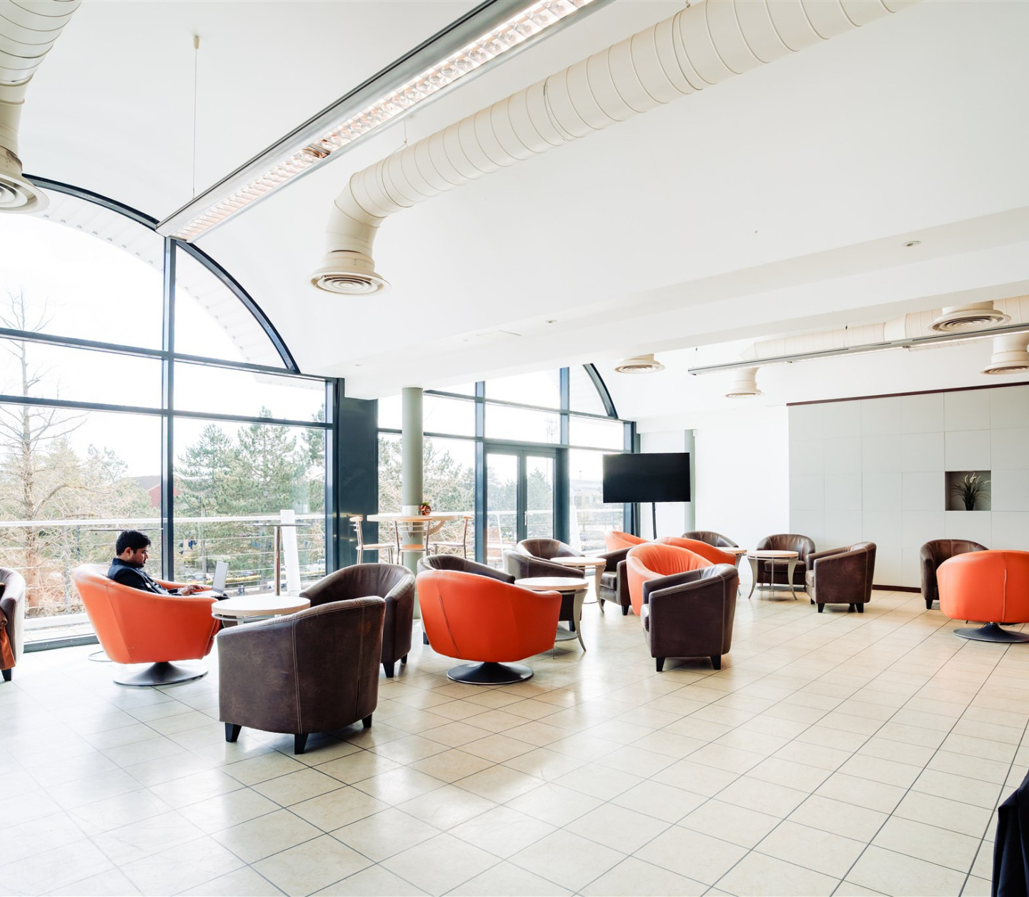 A spacious lounge area with large windows, featuring plush armchairs in orange and gray. A man is seated at a table, while another person is lounging on a chair nearby. The floor is tiled, and there is a modern design aesthetic.