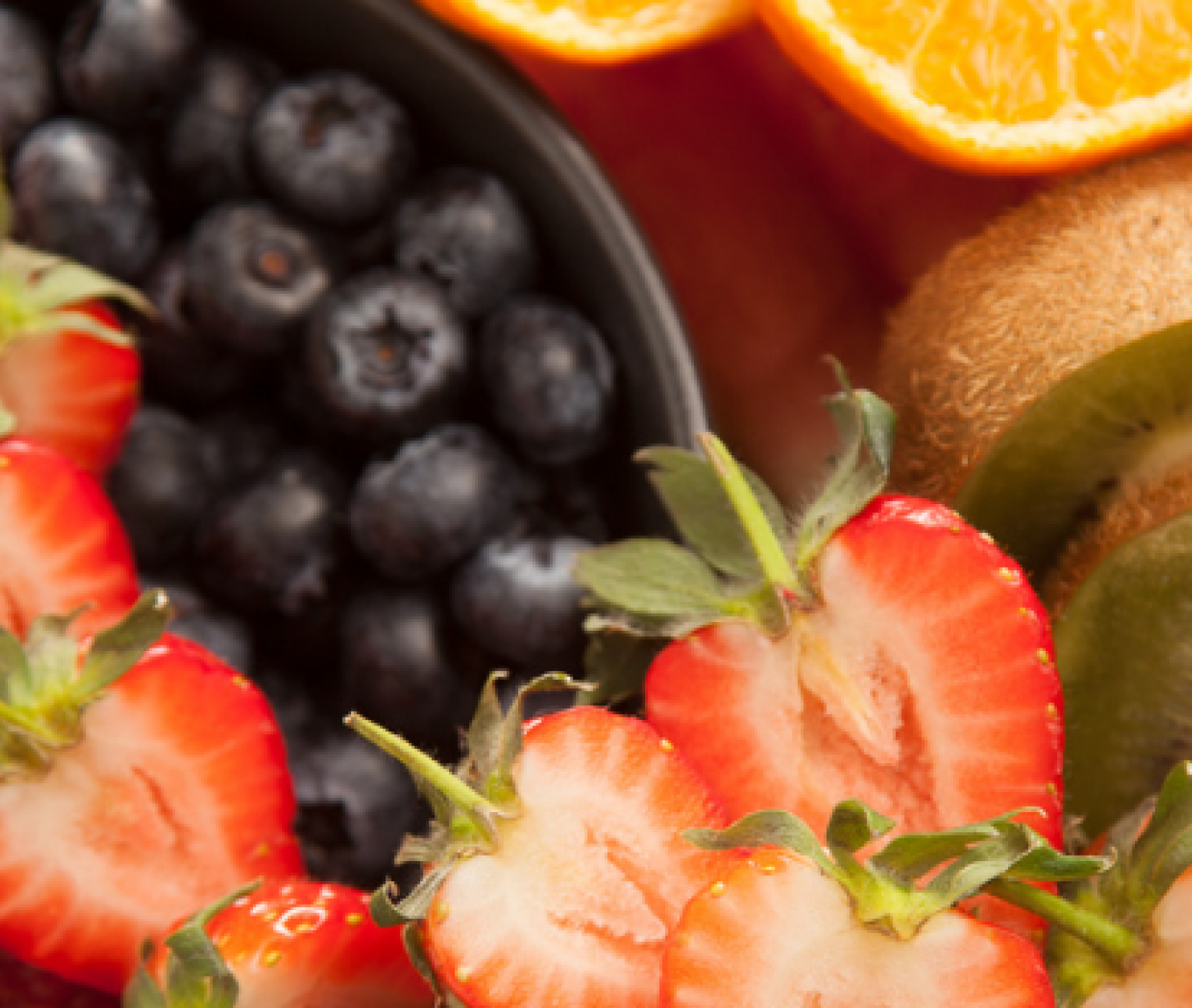 A colorful assortment of fresh fruits including strawberries, blueberries, raspberries, kiwi slices, and watermelon, arranged in a bowl with orange slices in the background.