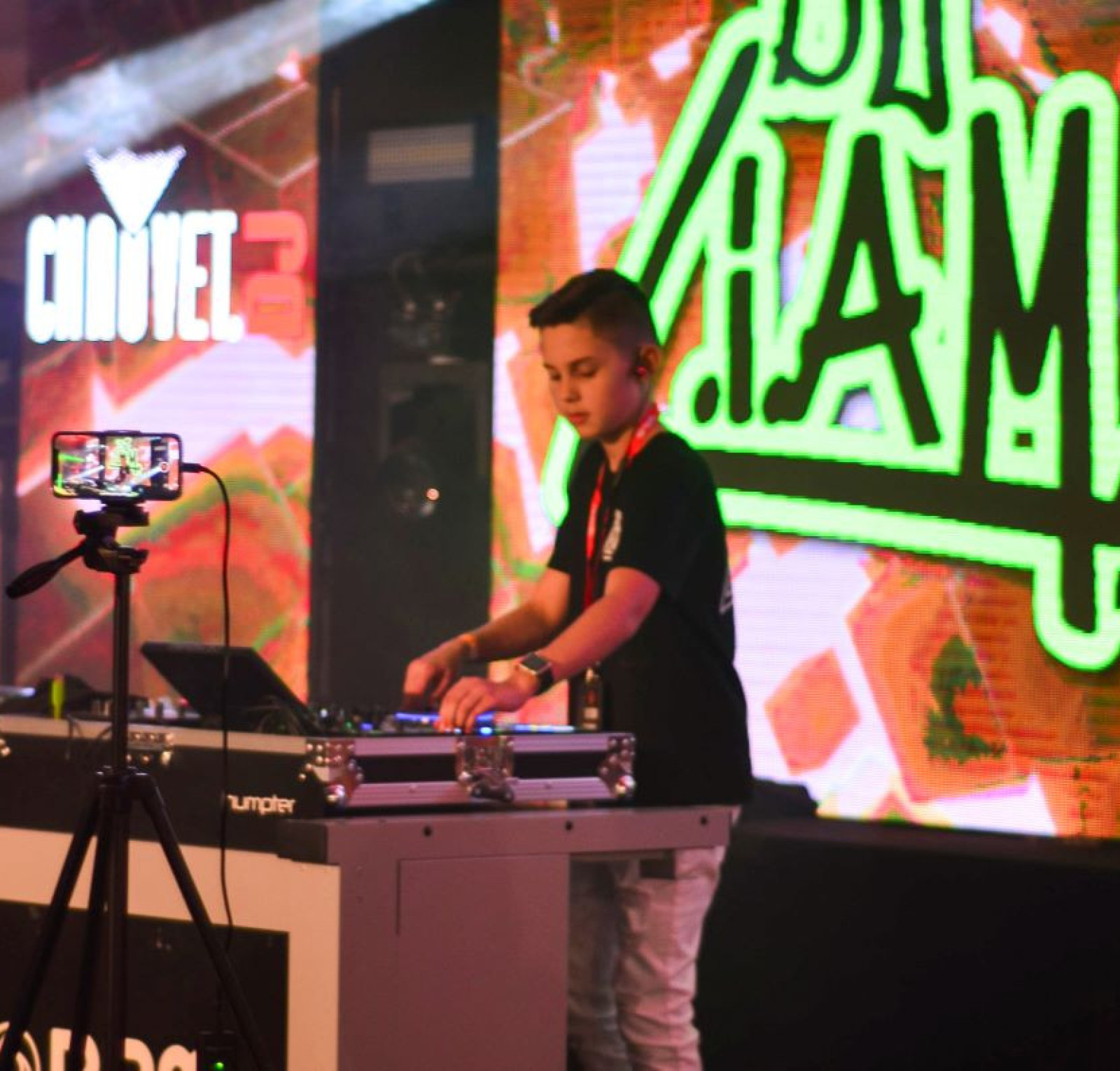 A young DJ stands behind a mixing console performing on stage. Colorful lights and large screens displaying the name 