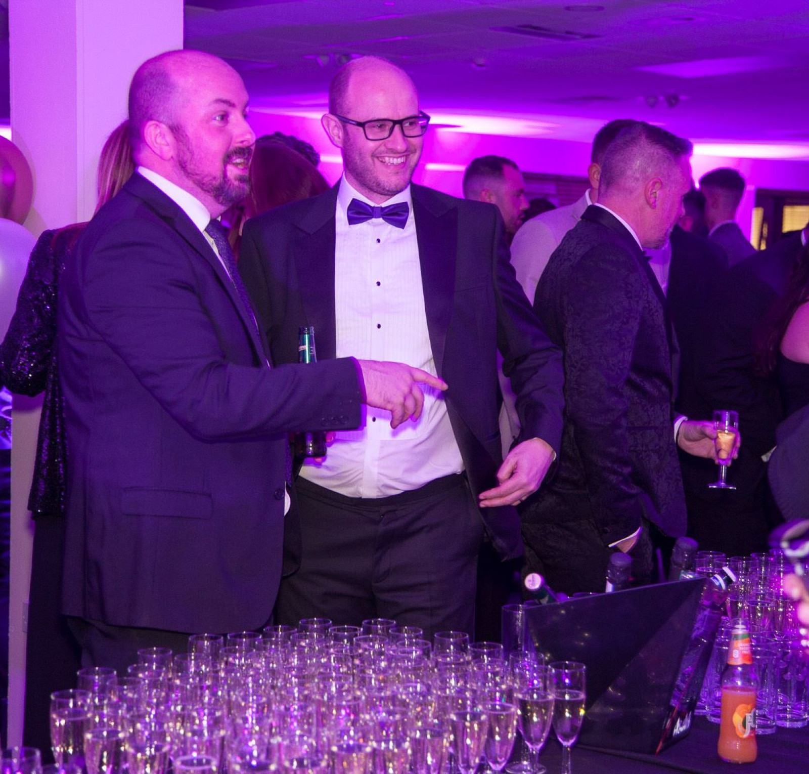 Two men in formal attire engage in conversation at a festive event. They are surrounded by guests and a table filled with glasses and drinks. The venue is decorated with purple lighting and balloons.